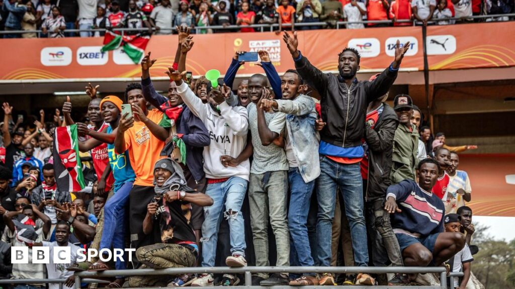 A large group of male Kenya fans pack on to a raised section obviously not designed for them at the Kasarani Stadium, with many clinging on to gain a vantage point. More fans can be seen in the stand behind them, which has orange CHAN branding and sponsors logos on it