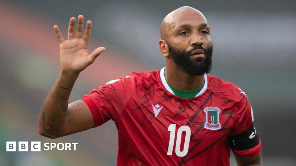 Emilio Nsue, wearing a red Equatorial Guinea shirt with a number 10 on the chest, holds his right hand aloft during a football game. The player has a bushy black beard and a neutral expression on his face