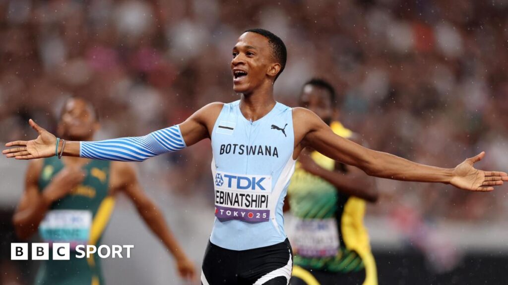 Collen Kebinatshipi seen from hips up, wearing a light blue Botswana running vest with a name bib on the front, black shorts and blue running sleeve covering part of his right arm, stretches out both arms in celebration after winning the 400m final at the World Athletics Championships in Tokyo. Blurred in the background are South African and Jamaican runners trailing in his wake and behind them a large crowd