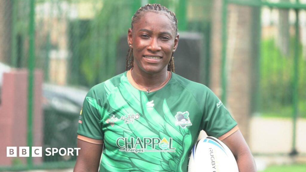 Rachel Iliya wears a short-sleeved green Nigeria rugby shirt while holding the rugby league oval ball tucked under her left arm. Blurred in the background can be seen a high chain-link fence with green supports spaced at intervals